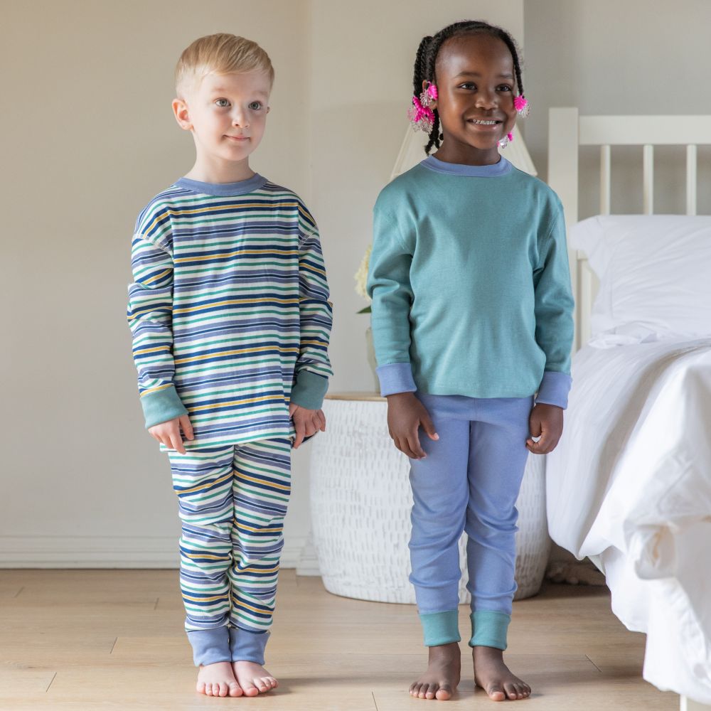Two children wearing pajamas standing in a bedroom. Eczema clothing cotton comfort kids pjs in multi stripe and green/blue