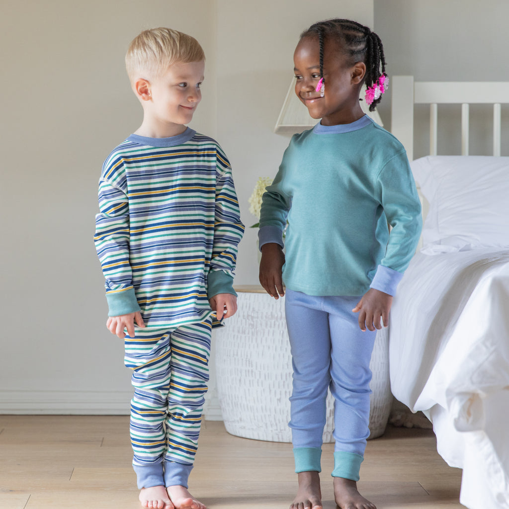 Two children wearing pajamas standing in a bedroom. Eczema clothing cotton comfort kids pjs in multi stripe and green/blue